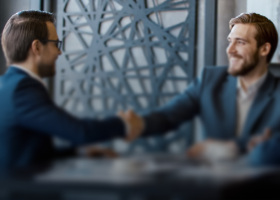 Two men in suits shaking hands at a meeting table in a modern office setting.