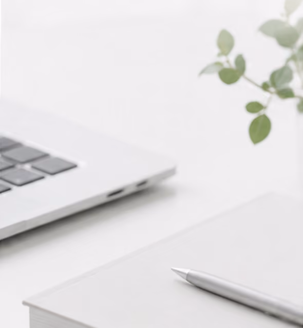 Silver pen resting on a closed laptop with another laptop keyboard partially visible and a small green plant.