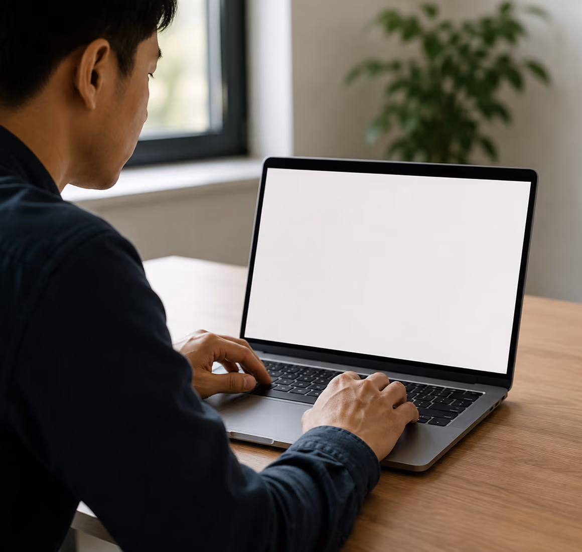 Man typing on a laptop with a blank white screen at a wooden desk near a window and plant.