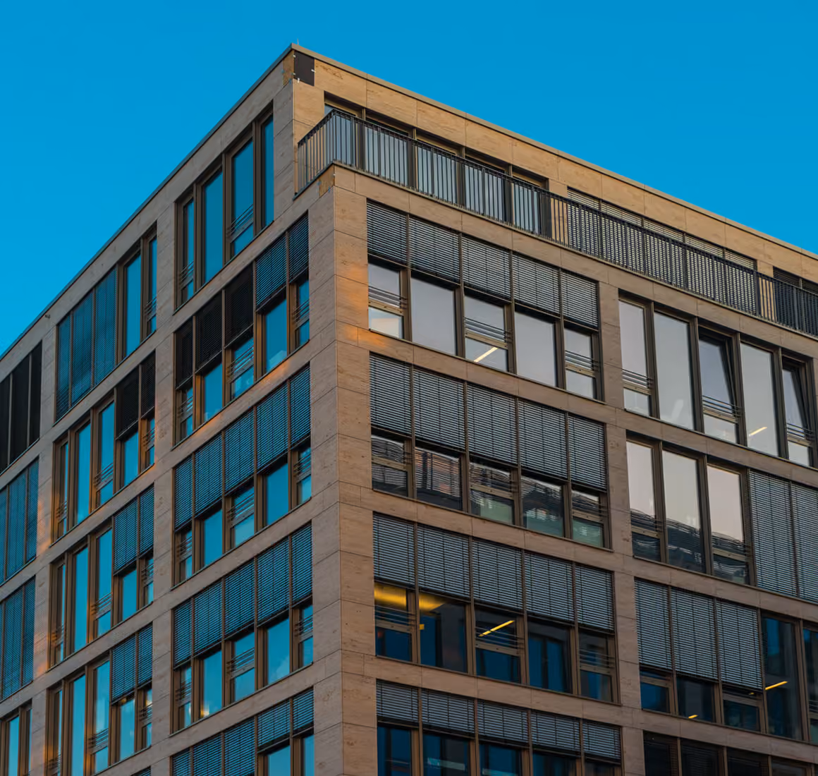 Modern office building corner with glass windows and metal blinds against a clear blue sky.